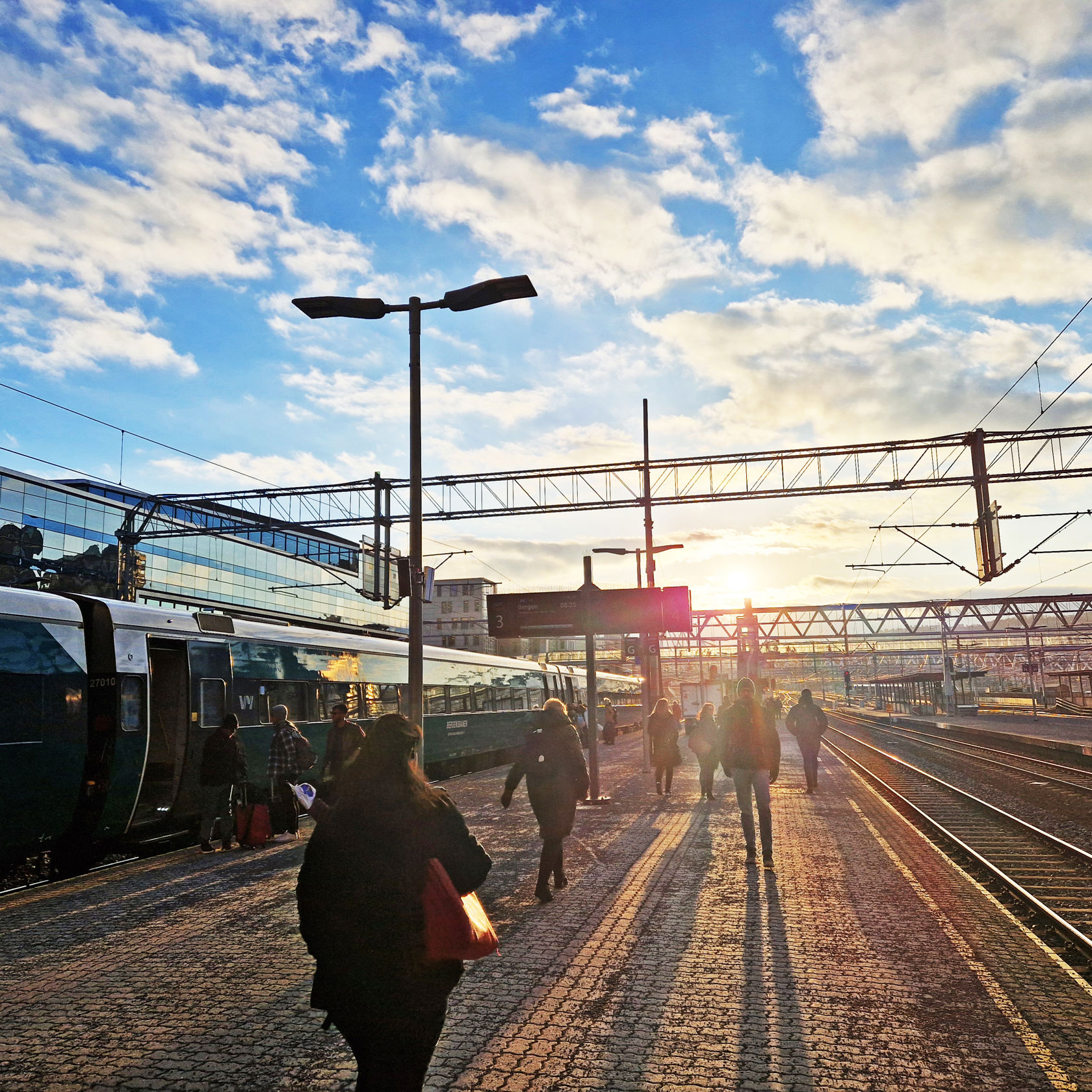 Lav sol over perrongene på Oslo S. Folk går på plattformen mot sola. Et tog står på ett av sporene.