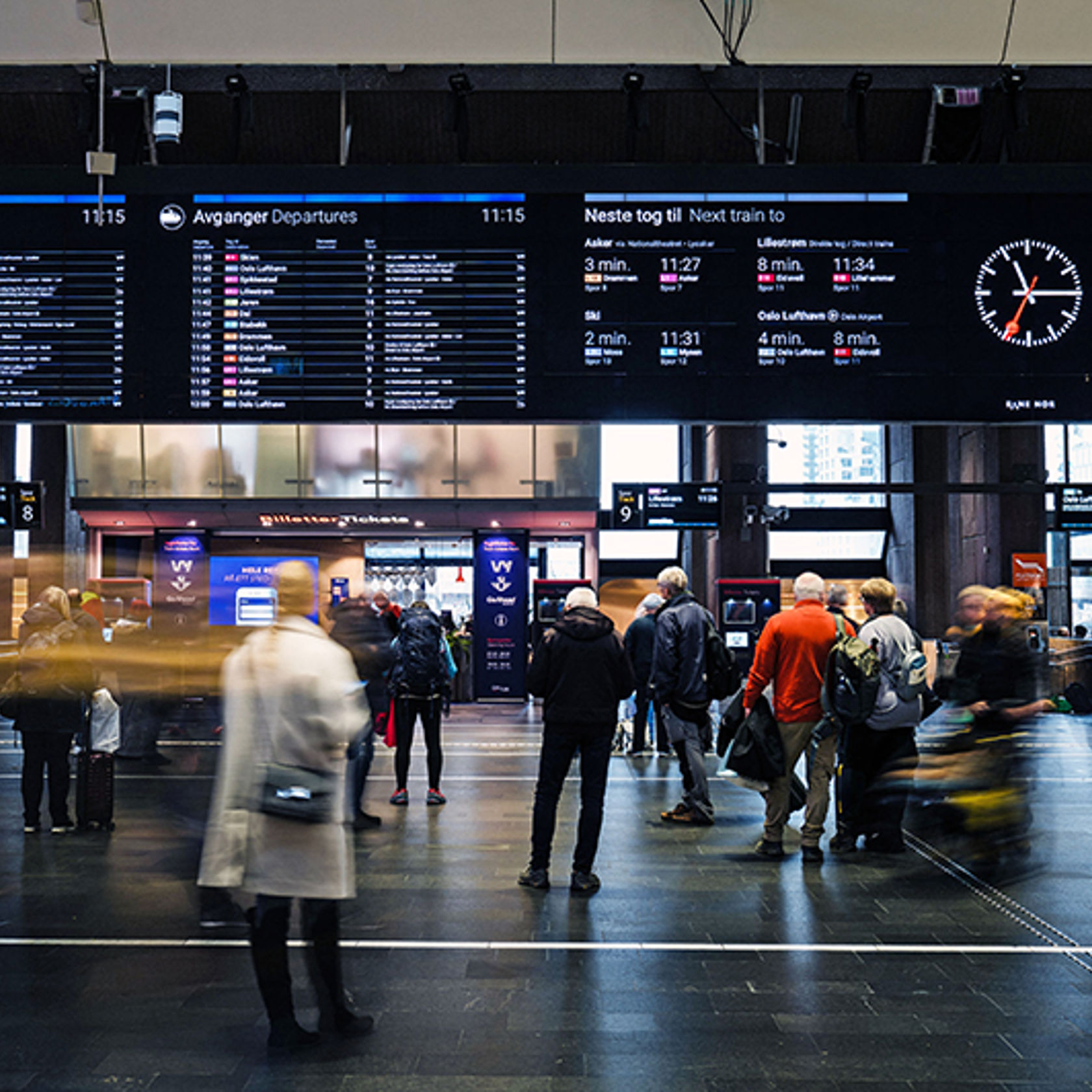 Folk som står under en infoskjerm på Oslo S som viser togtidene.