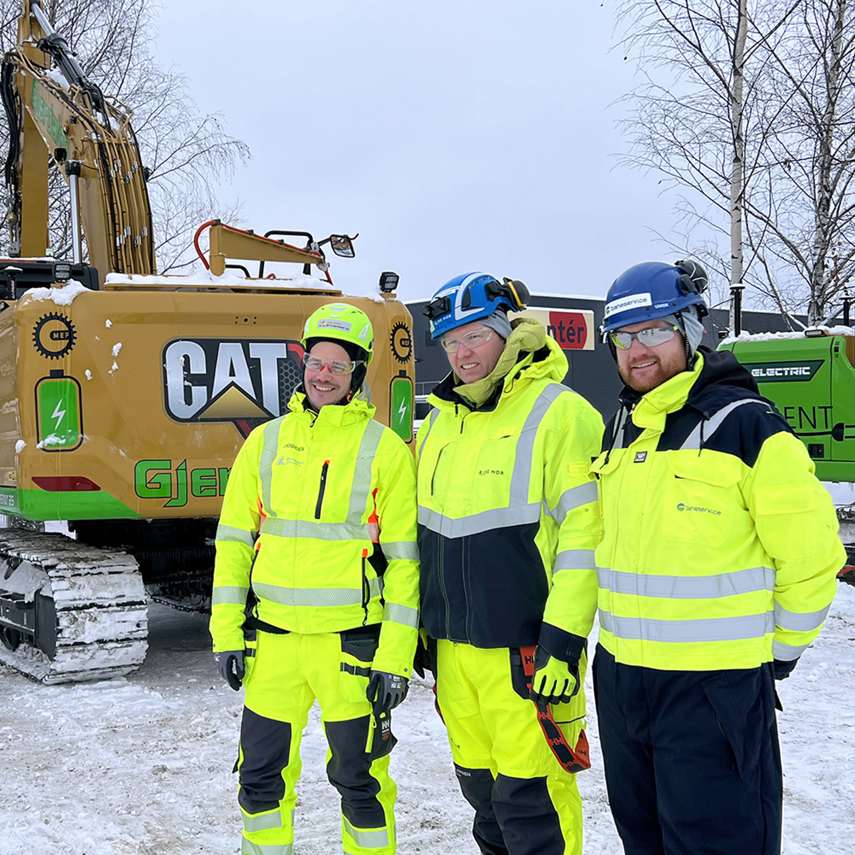 Personene i anleggsutstyr foran to gravemaskiner tilkoblet ladekabler. 