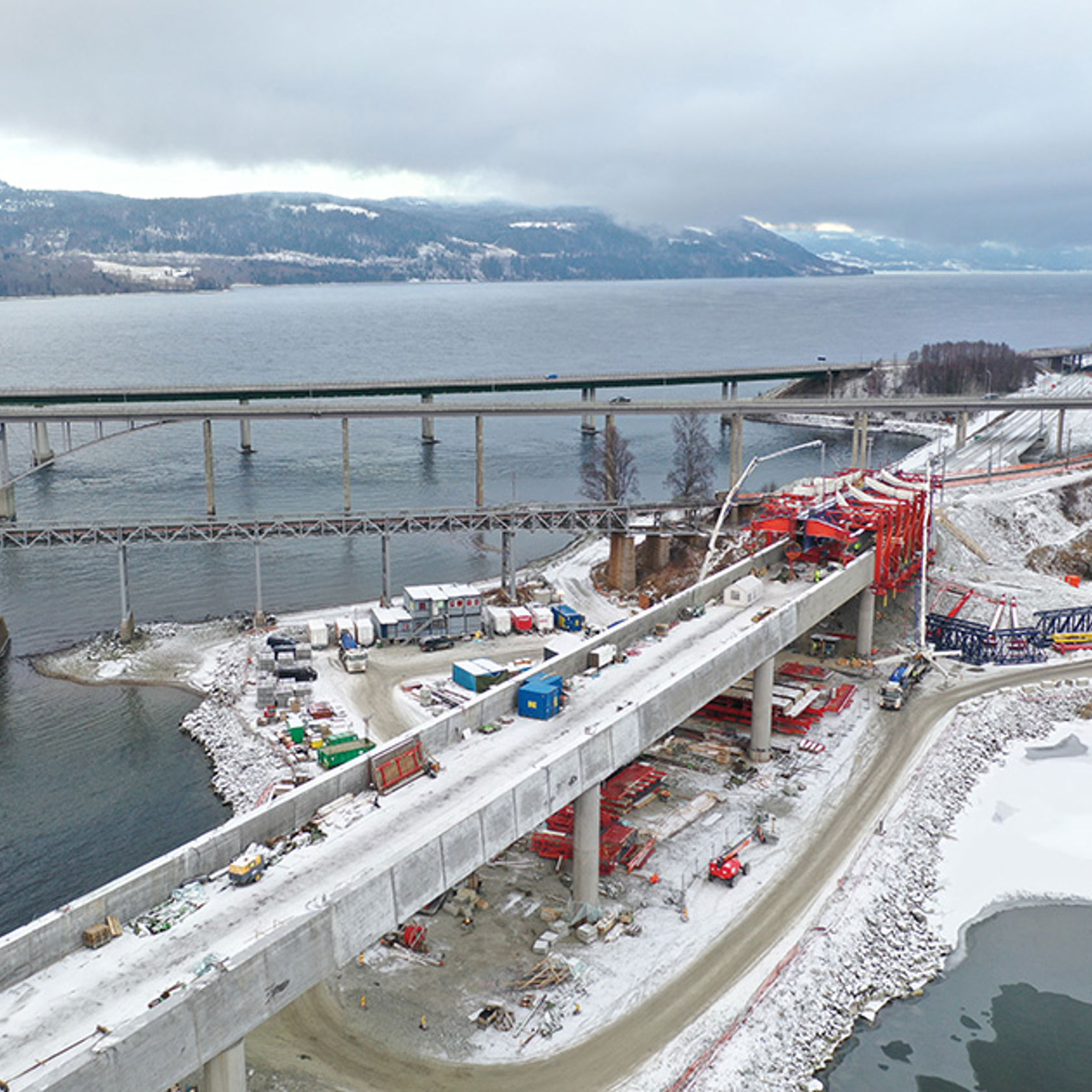 Dronebilde av brua, forskalingsvogna og betongbiler/pumper mens støpen pågår. I bakgrunnen Mjøsa og de andre bruene på Minnesund.