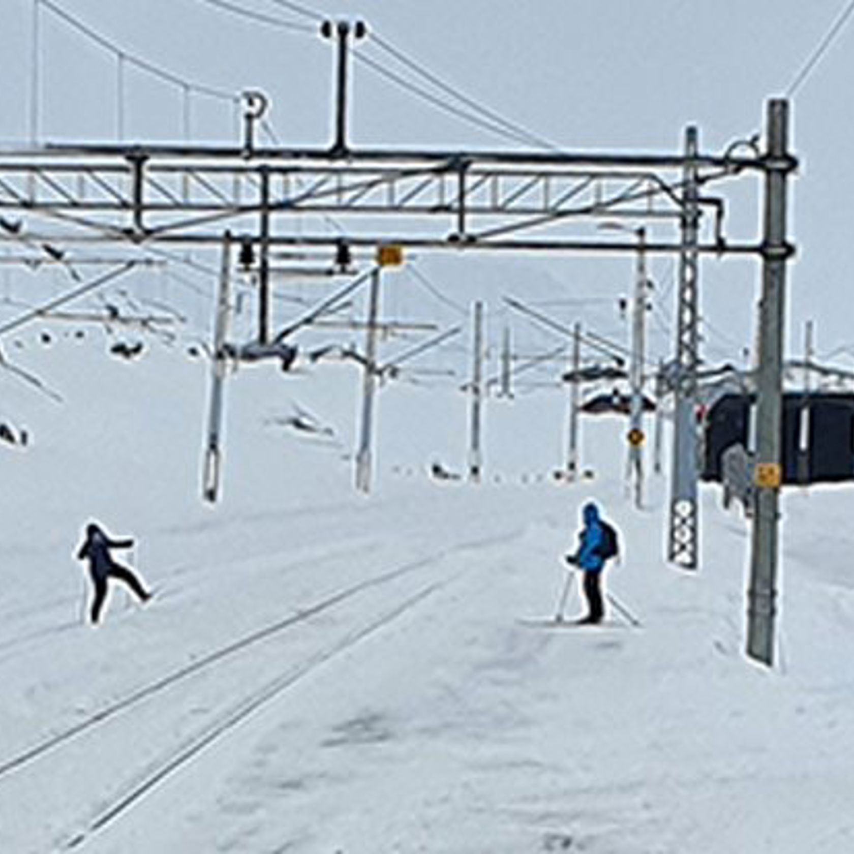 To skigåere tråkker over jernbanens spor inne på en snødekt Finse stasjon.