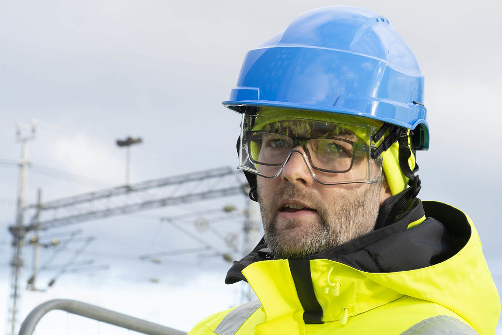 Personkledd i gult vernetøy og blå hjelm. I bakgrunnen synes en jernbanemast.