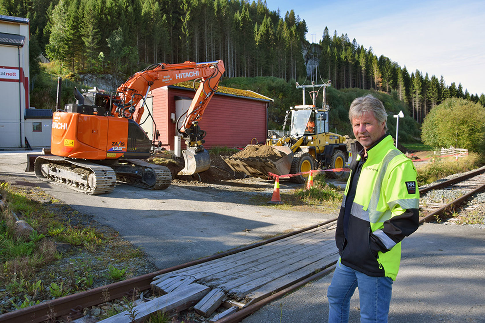 Mann i vernetøy står foran et anleggsområde. Bak han er en gravemaskin i ferd med å grave i jorda. 