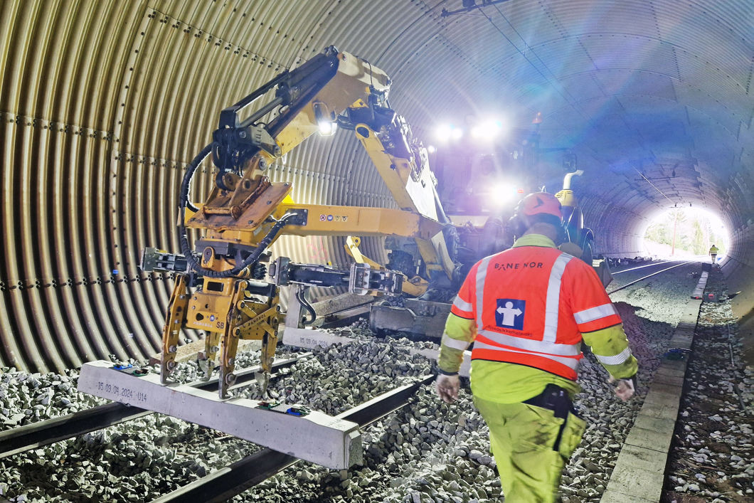 En arbeidsmaskin jobber på skinnegangen inne i en tunnel. En mann i verneklær går mot maskina.