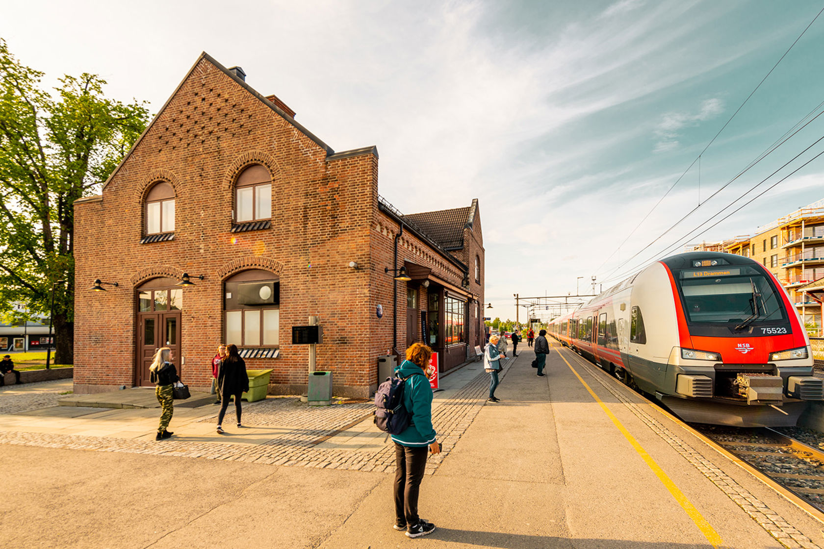 Illustrasjonsfoto fra Jessheim stasjon med stasjonsbygning, passasjerer og tog ved plattform.