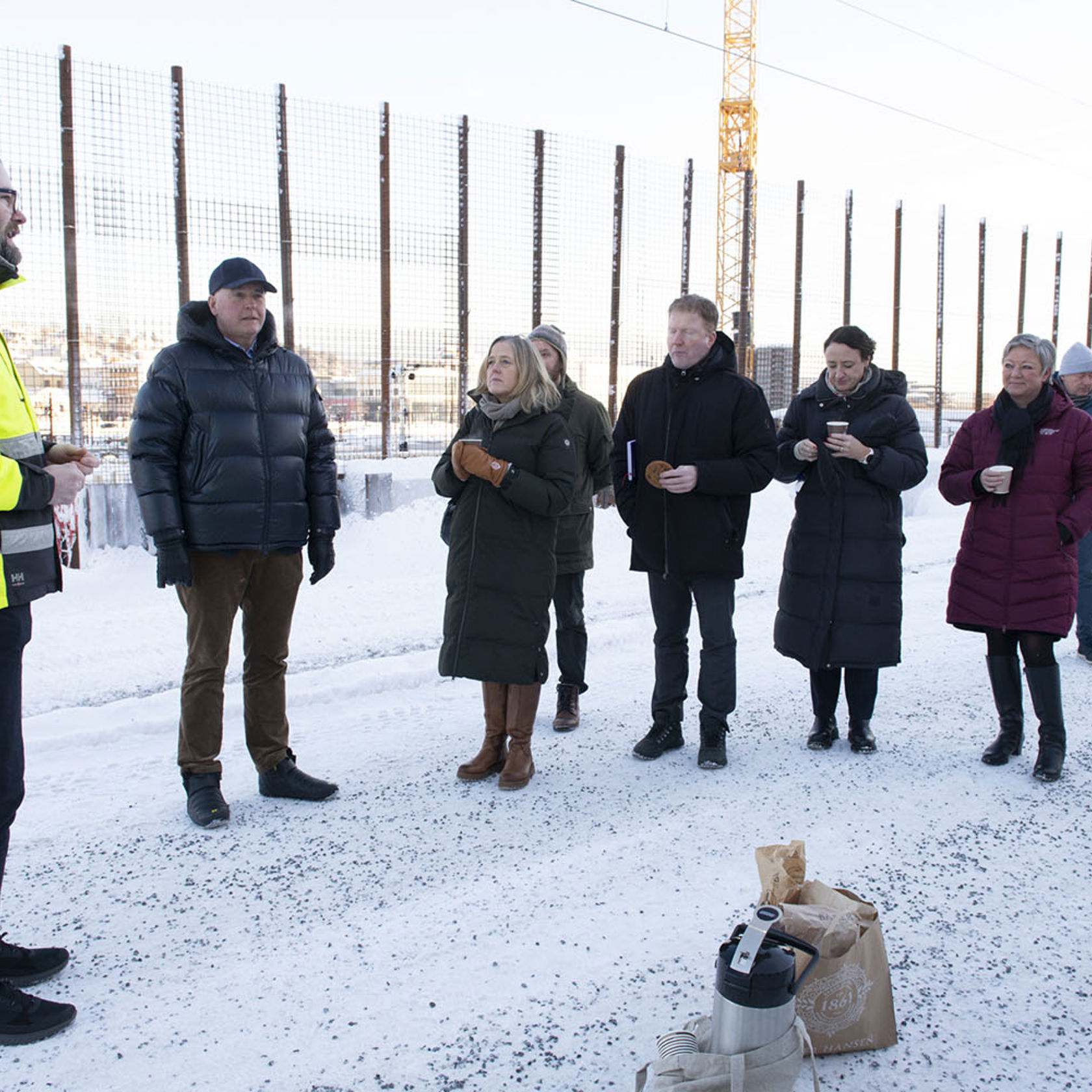 Gruppe mennesker med kaffe, gjerde og byggekran, vinter.