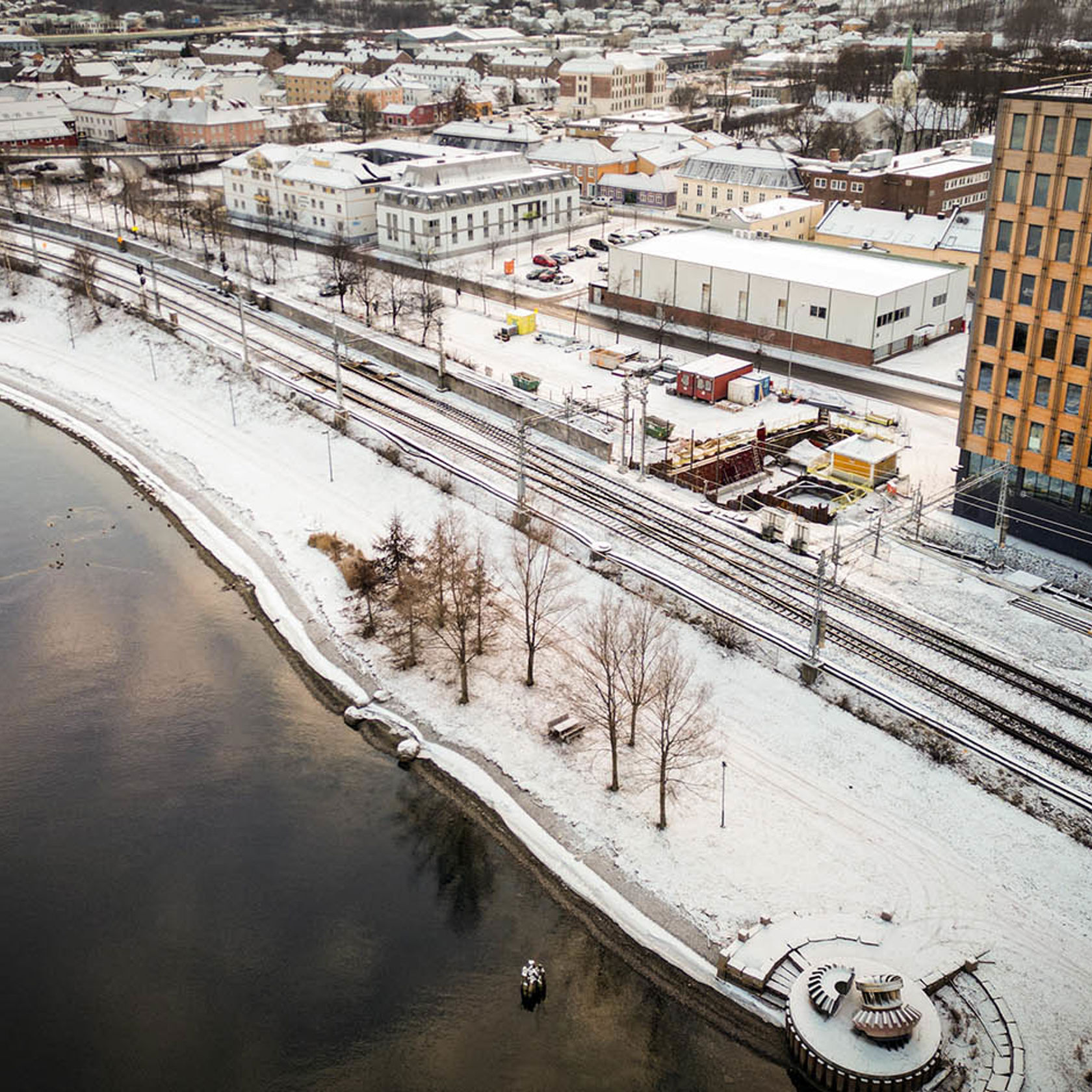 Sporområde og elvepromeade sett fra luften. Snø på bakken. Synlig anleggsområdet langs sporene.