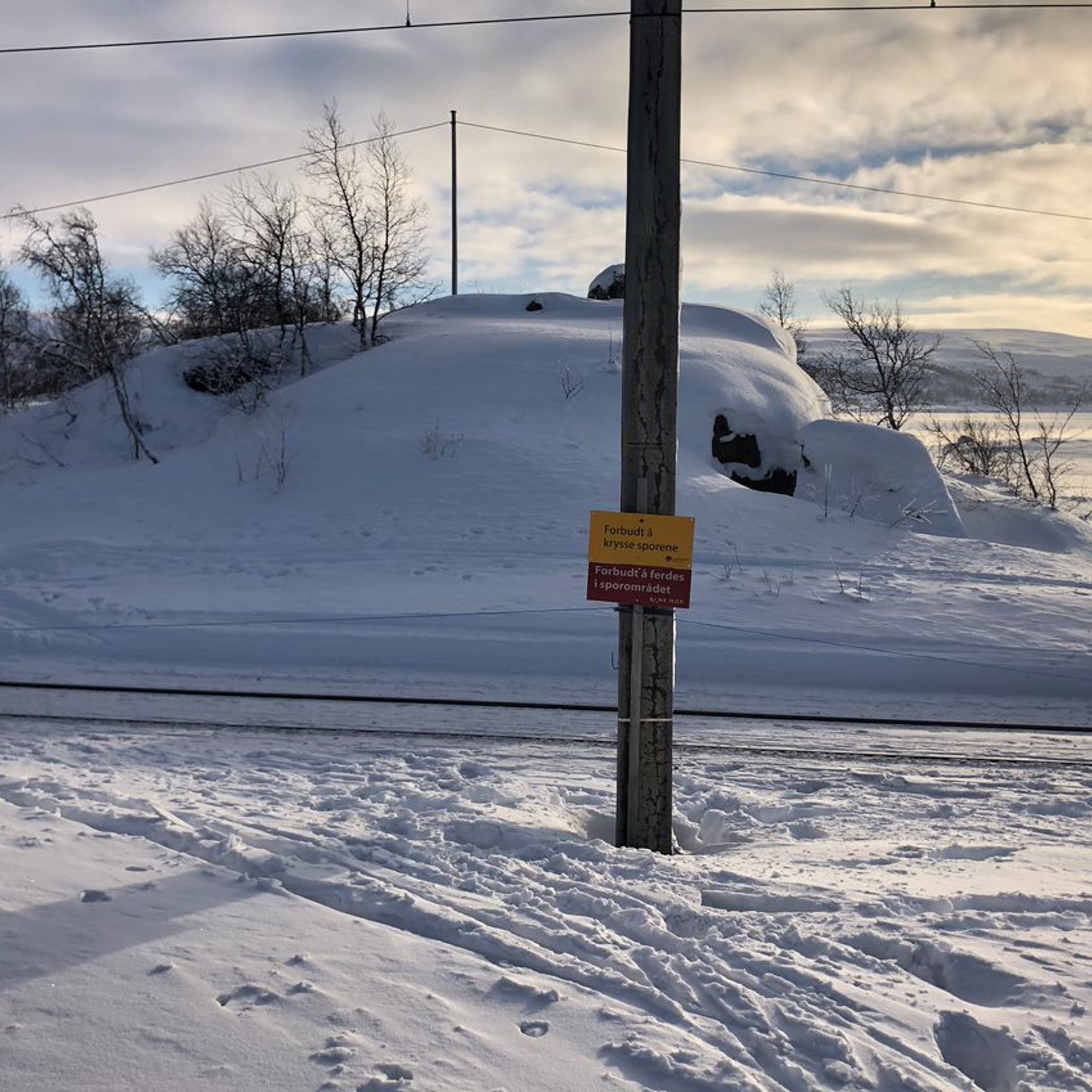 I et snødekt terreng står det en stolpe med advarsel om å krysse jernbanen. Ved siden av går skispor som krysser jernbanen.