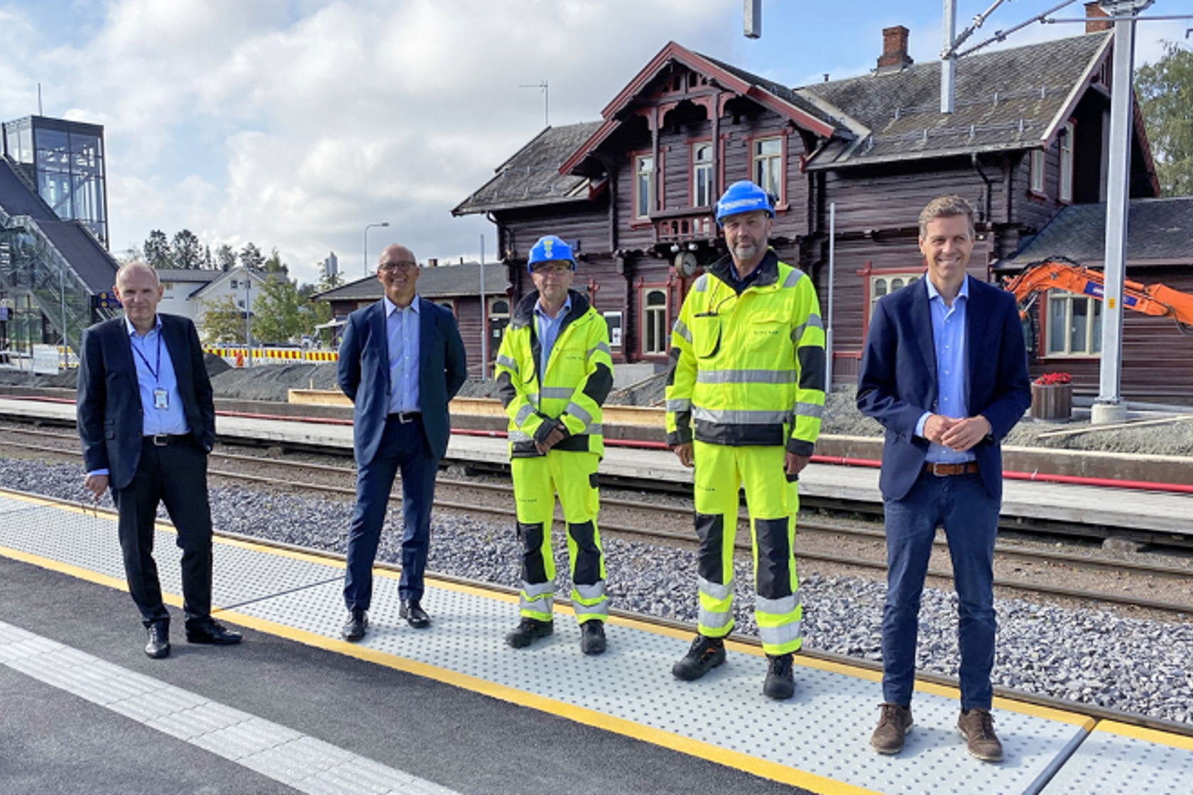 Bilde av tre menn i dress og to i arbeidsklær som står på en togperrong.