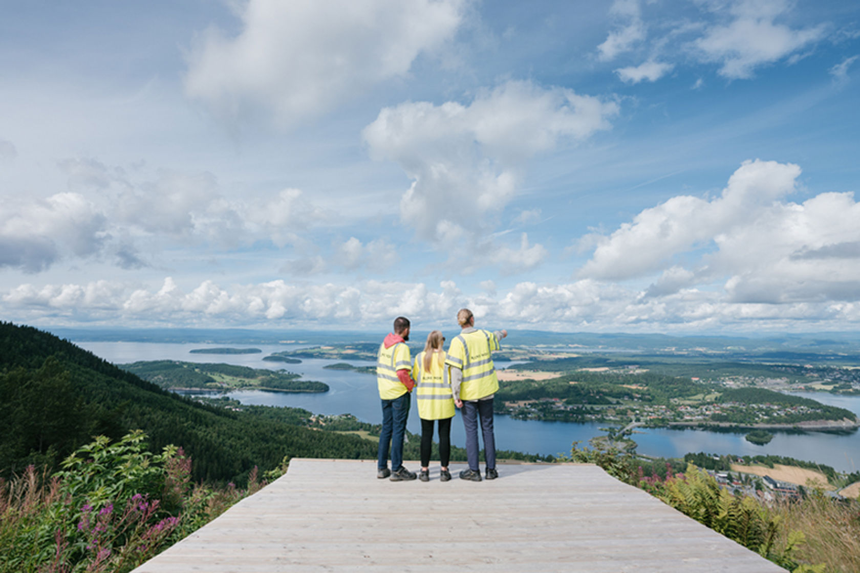 Tre personer med gule Bane NOR-vester står og skuer utover et landskap med sjø og flatt innland.