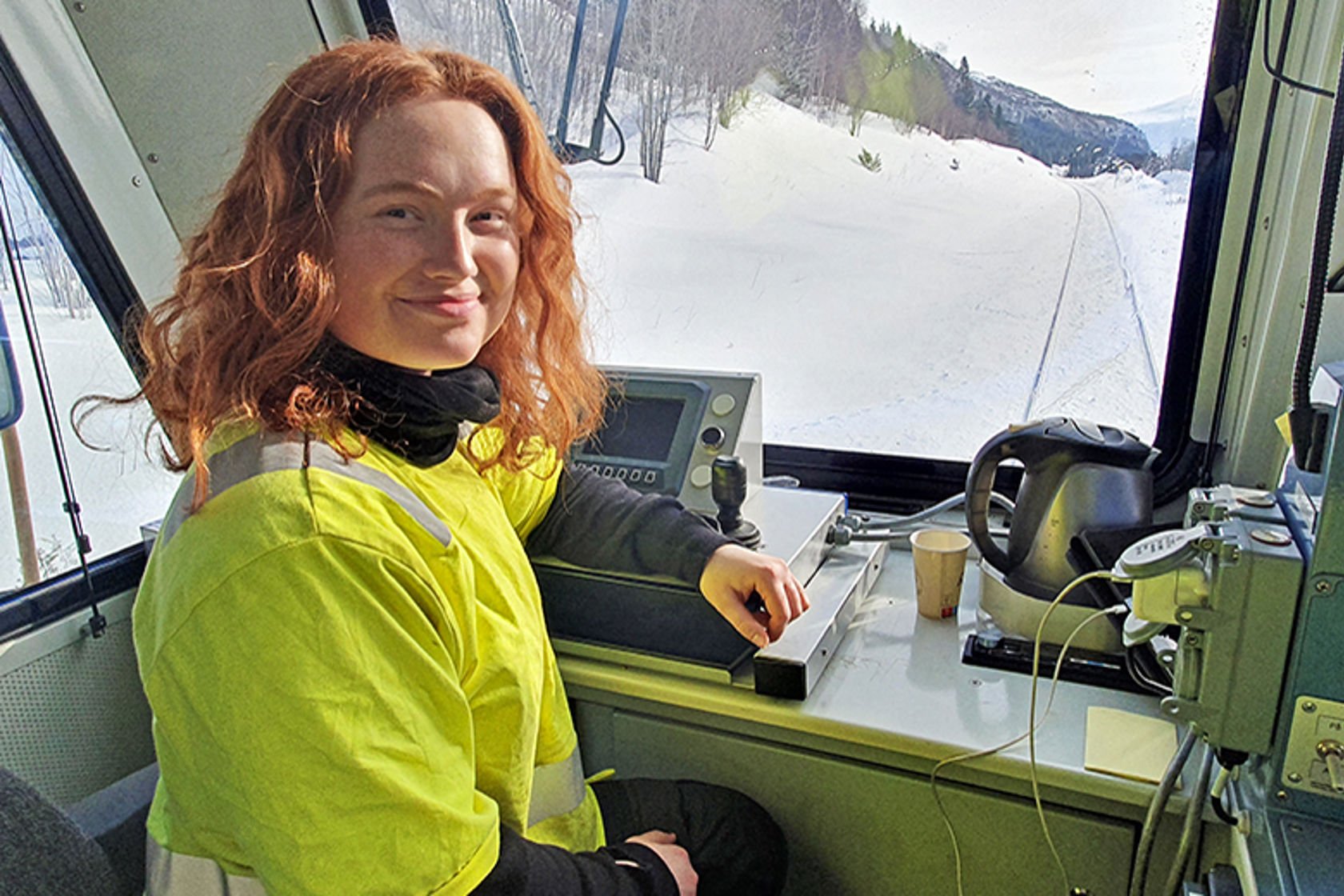 En ung kvinne i arbeidstøy sitter ved spakene i et arbeidstog på jernbanen.