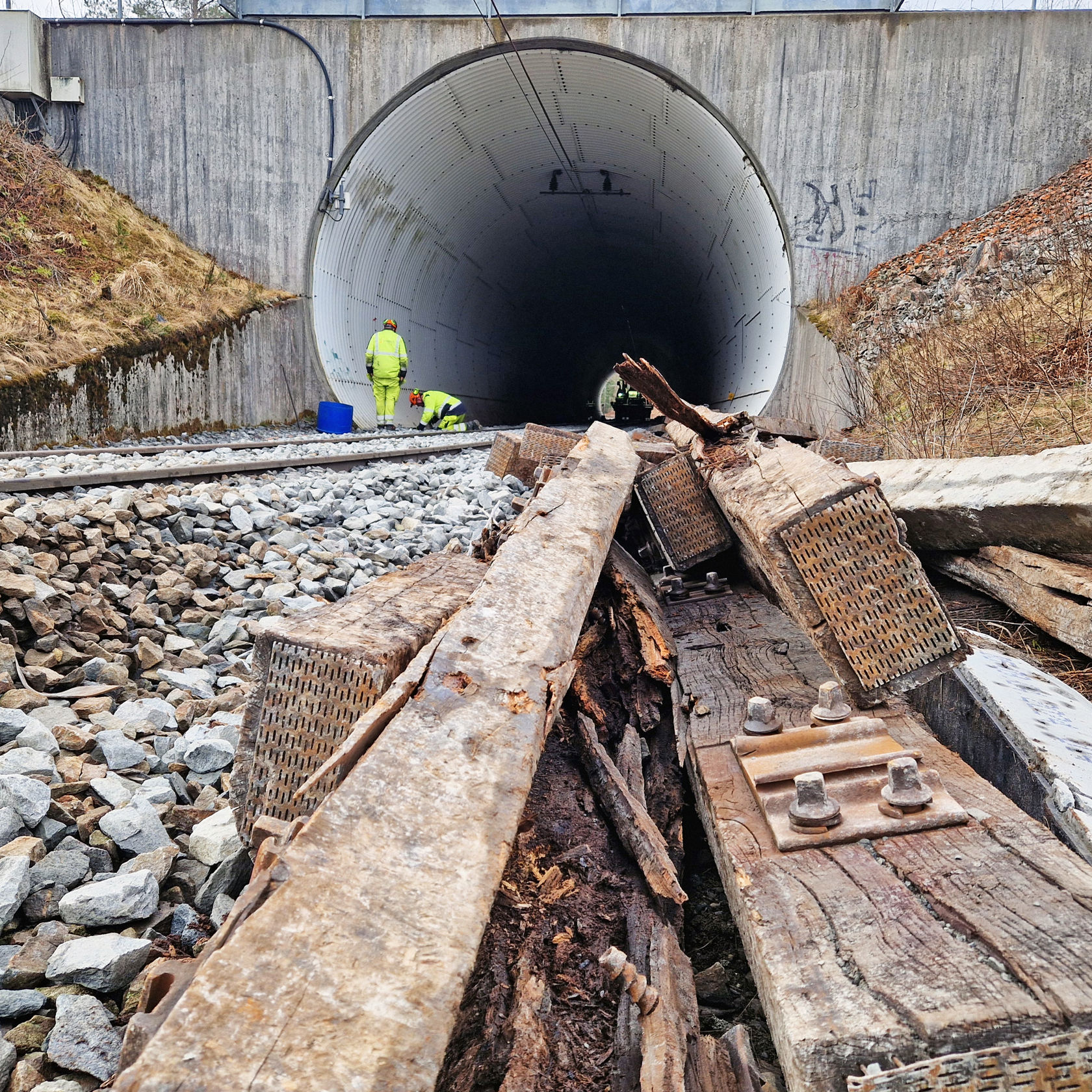 Noen gamle, råtne tresviller i forgrunn, jernbanespor som går inn i en tunnel i bakgrunn. 