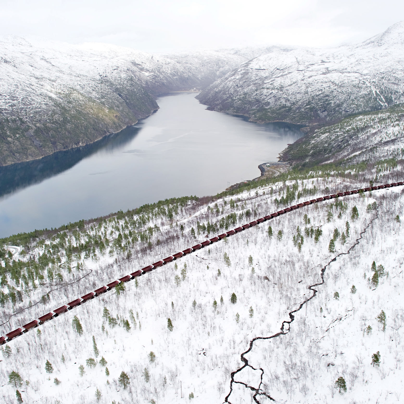 Bilde tatt fra lufta av et langt malmtog som går gjennom et hvit fjellandsskap med en innsjø.