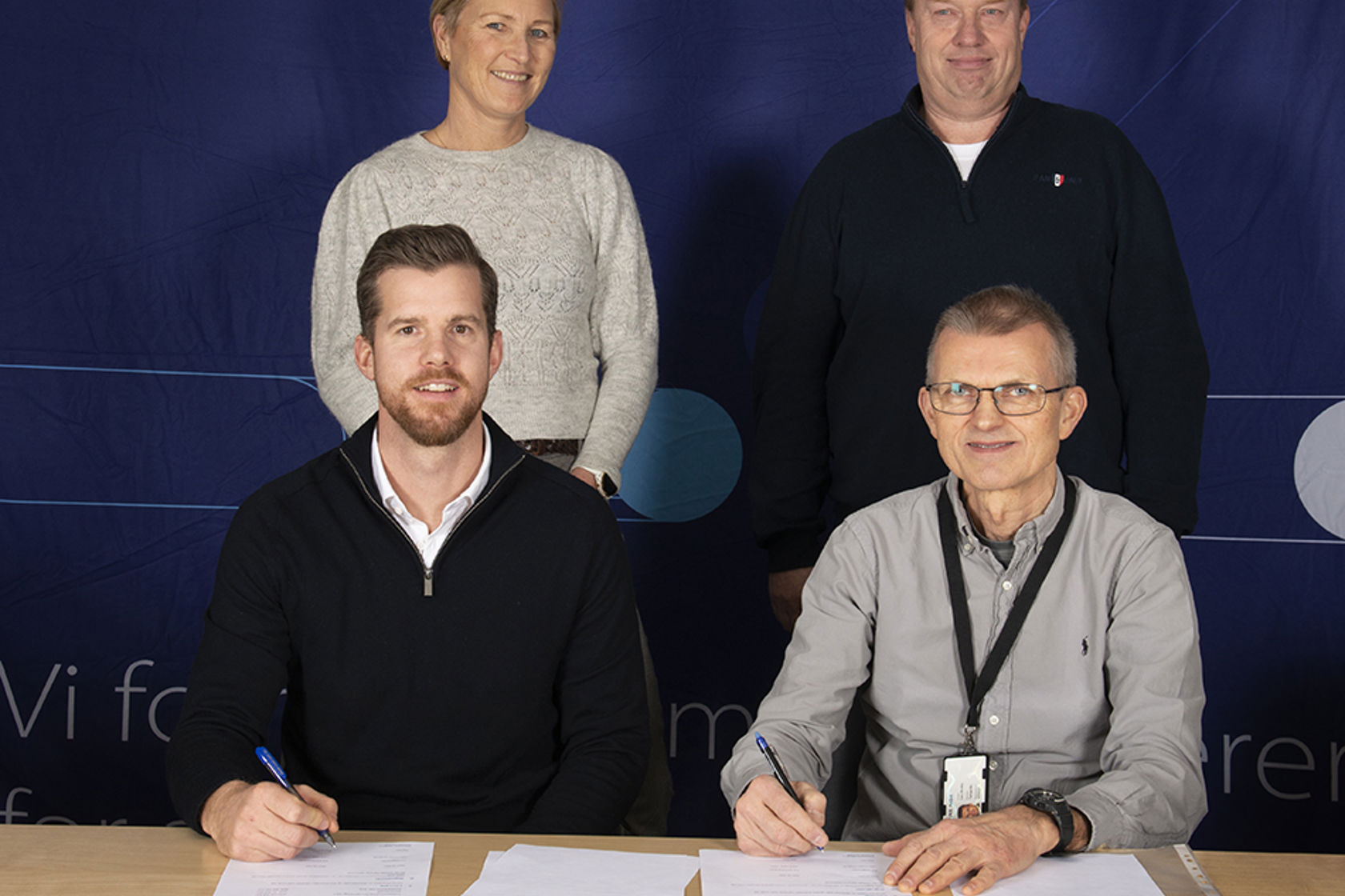 To menn signerer kontrakt, en kvinne og mann står bak. Foran på bordet står det en bløtkake