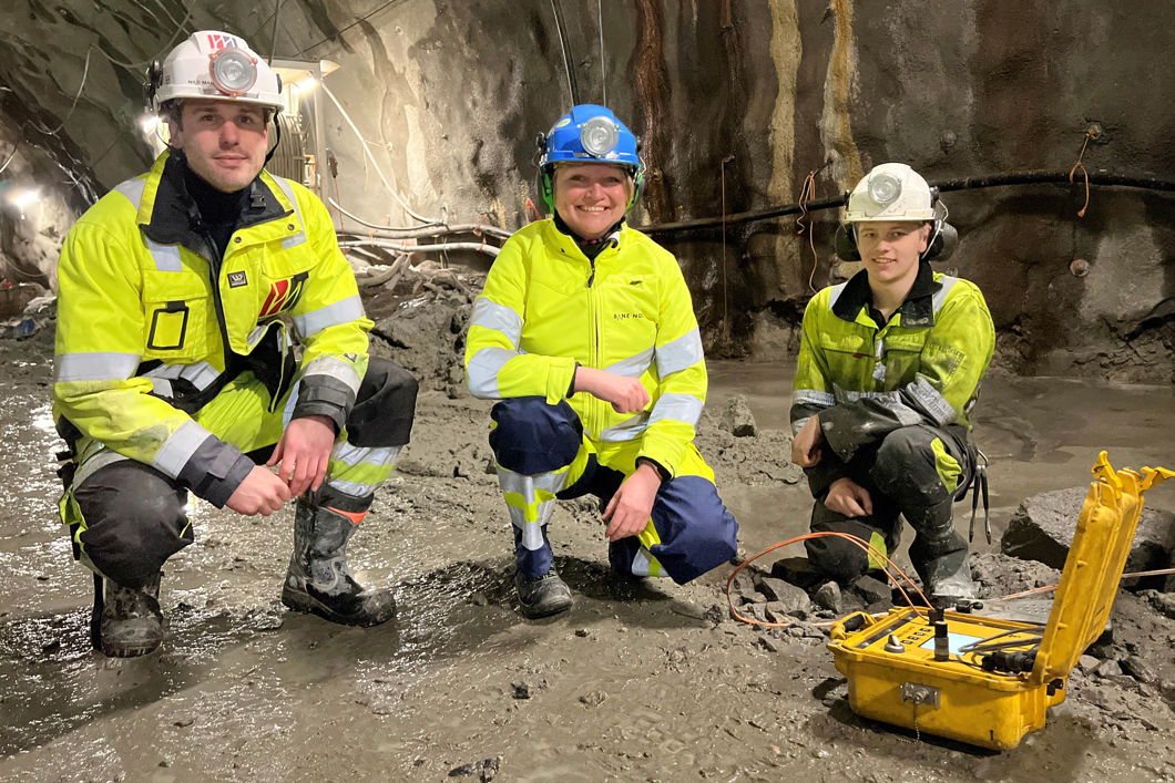 To menn og en kvinne står på huk iført vernetøy i tunnel