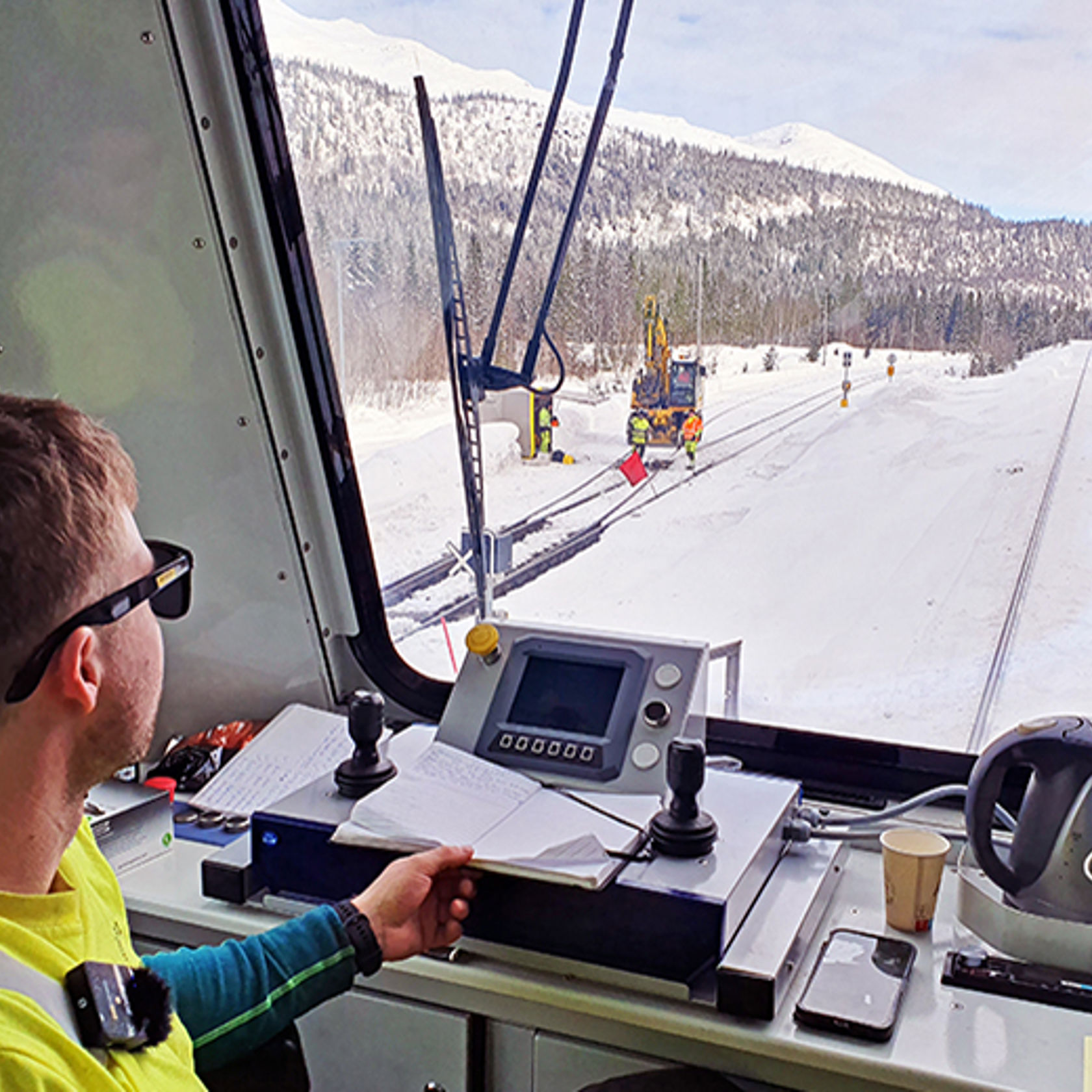 Mann sitter ved spakene i et arbeidstog med utsikt mot skog og snødekte fjell.