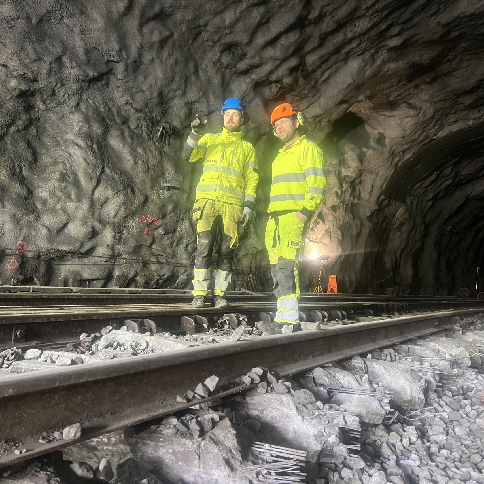 To arbeidsmenn i gule klær står på jernbaneskinner i en tunnel. En peker innover i tunnelen.